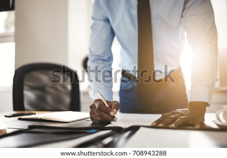 Young African entrepreneur wearing a shirt and tie leaning on his desk in an office signing documents 