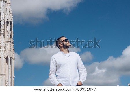 Photo of a young handsome man with sunglasses looking up to the sunny sky, with blue cloudy sky in the background. Tourist in Milan, Italy