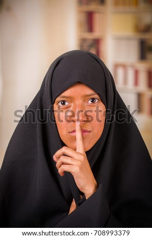 Portrait of a beautiful muslim girl wearing a hijab, and doing a silence sign with her hand, in a blurred background