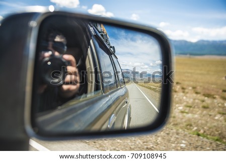 Man or woman taking photo with professional camera in a car side mirror with road and natural mountain background. asphalt road in perspective. blue sky with clouds