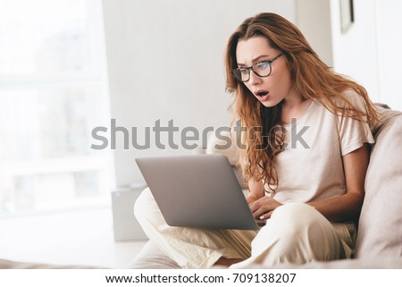 Image of young shocked lady sitting on sofa indoors. Looking aside using laptop computer.