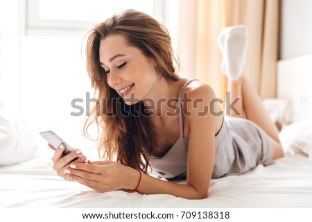 Image of young happy emotional pretty lady lies on bed indoors. Looking aside chatting by phone.