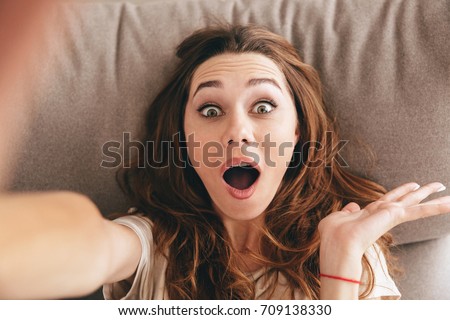 Image of young amazing shocked emotional pretty lady lies on sofa indoors. Looking camera make selfie.