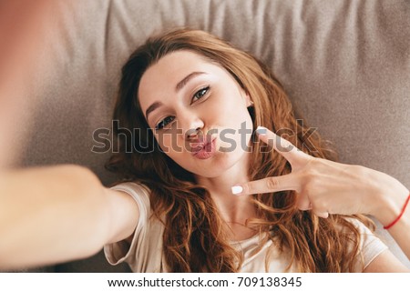 Image of young amazing emotional pretty lady lies on sofa indoors. Looking camera make selfie make peace gesture.