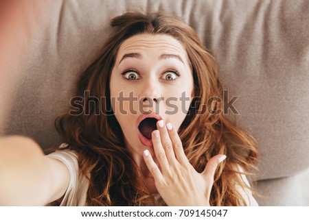 Image of young amazing shocked emotional pretty lady lies on sofa indoors. Looking camera make selfie.