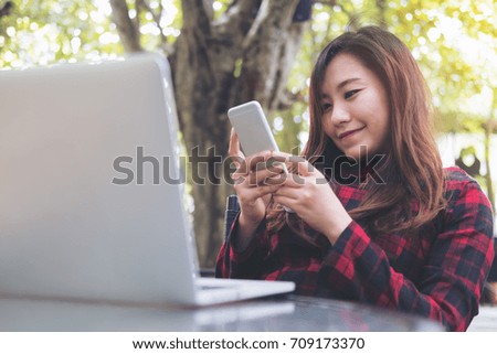 Closeup image of a beautiful Asian business woman using and looking at smart phone with laptop on glass table at outdoor with green nature background