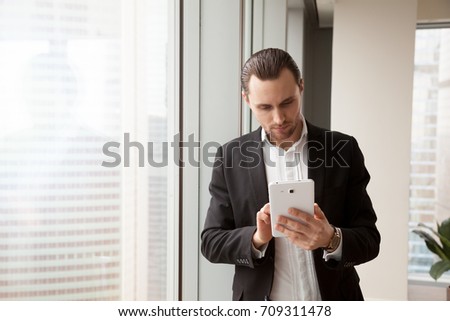 Smart young well-groomed handsome businessman in expensive dress suit is standing next to window in modern office and looking for information on his electronic tablet, marketing or investing concept.