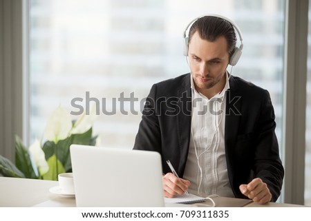 Thoughtful businessman in headphones taking notes in front of laptop at workplace. Young manager participating in online meeting or conference, remote job interview, learning foreign languages.