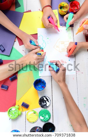 Markers in male and female hands draw on white paper with little drawings. Artists wooden table with paints and colored paper. Art and idea concept. Hands hold colorful markers and draw