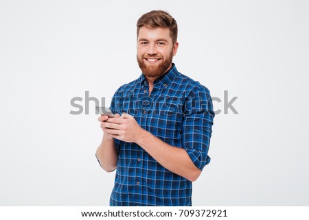 Pleased bearded man in checkered shirt using smartphone and looking at the camera over gray background