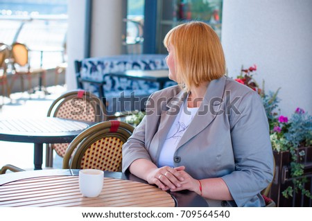 A woman of size plus a US or European look sitting in a restaurant drinks coffee in a good mood. A young girl with excess weight, stylishly dressed in the center of the city is waiting for a friend
