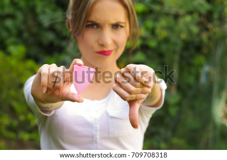 Close up of a young woman pointing in front of her a menstrual cup in one hand, Gynecology concept, ith her thums down rejecting the use of the mentrual cup, in a blurred background