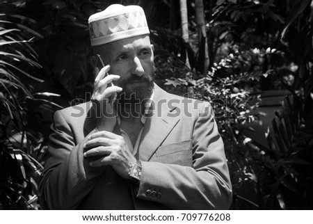 Black and white portrait of a bearded man wearing a jacket and an ornate taqiyah talking on a cellphone outdoor