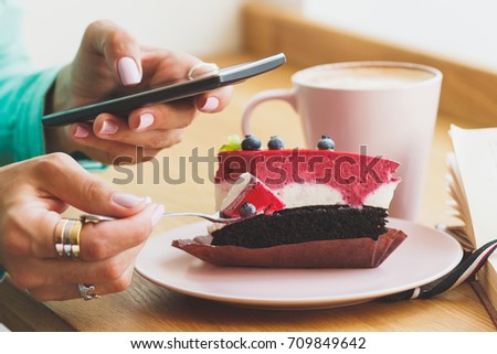 Woman takes a picture of cup with cappuccino and a piece of dessert on the plate, soft focus background
