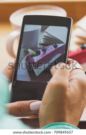 Woman takes a picture of cup with cappuccino and a piece of dessert on the plate, soft focus background