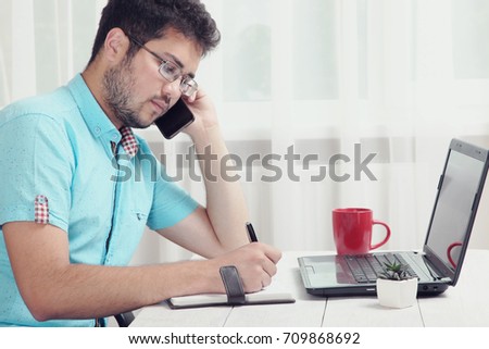 A guy talking on the phone and writing in a notebook sitting at the table. 