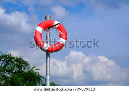 Red swim tube for lifeguard rescue hanging on the metal pole at the lake with green tree and blue sky background 