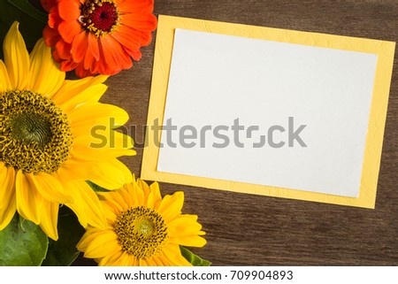 White blank greeting card with beautiful yellow and red flowers on the brown wooden table. Empty place for a text. Top view.