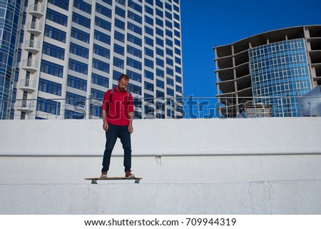 Young bearded man riding on skateboard, hipster with longboard in red 
shirt and blue jeans urban background 