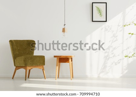 Handmade lamp above wooden table in living room with comfortable green chair and poster of leaf on white wall