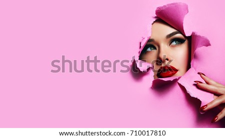 the face of a young beautiful girl with a bright make-up and with plump red lips peeks into a hole in pink paper. Nails with bright red lacquer.Lipstick,cosmetics,makeup, fashion, beauty,beauty salon. Royalty-Free Stock Photo #710017810