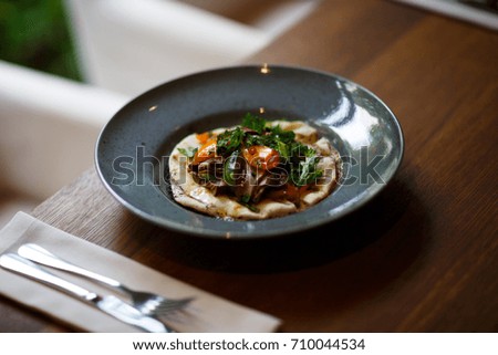 A close-up picture of a blue round plate full of tasty dish next to a dinner service on a white table napkin on a dark wooden background. Delicious expensive food. Restaurant concept.