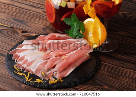 A glass jug of refreshing fruit beverage with slices of apples and orange, a plate of thinly sliced prosciutto and fresh green leaves of mint on a wooden brown background.