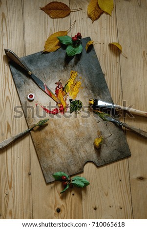 Wooden palette with oil paints yellow red, brushes and palette knife over wooden floor. With autumn decoration yellow leaves and red berries. Top view with space. Art concept. Atmospheric day light