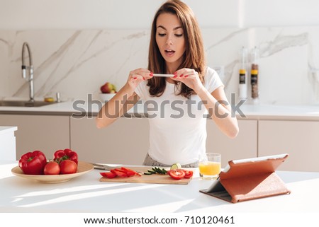 Happy smiling woman taking a picture of vegetable slices on a wooden board with mobile phone while standing at the kitchen