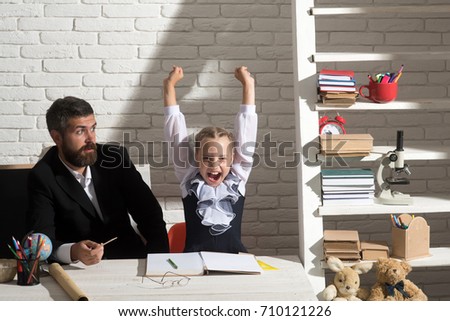 Daughter and father in classroom on white brick background. Education and family relationship concept. Schoolgirl and dad with cheerful and surprised faces. Man and girl by desk with school supplies