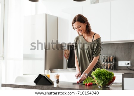 Smiling casual woman cuts vegetable by the table on kitchen and using tablet computer