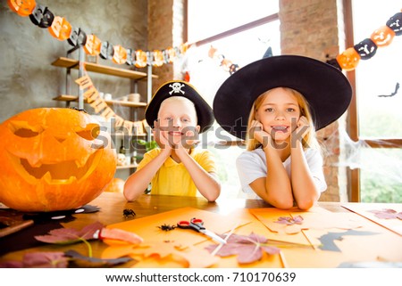 Close up cropped photo portrait of two small kids at halloween party - blond small witch and pirate, bonding, holding heads with arms, cheerful toothy smiles, indoors at home, loft style decorated
