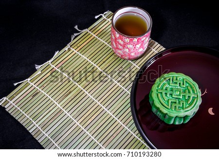 A jade moon cake on a dark plate, a tea cup with pink floral patterns, on a bamboo mat against a black background. Space for words available.