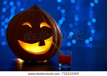 Close up of red smiling pumpkin with one tooth and big eyes, decoration prepared for Halloween smiling and drinking cocktail at party. Dark blue background with lights. Autumn holiday.