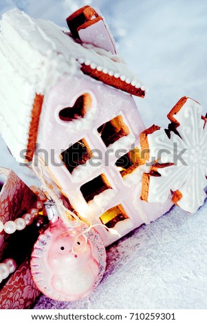Magical winter christmas picture. Gingerbread house with pink icing with light shining in the windows under snow falling and new year toys and decorations in paper box with cookies.