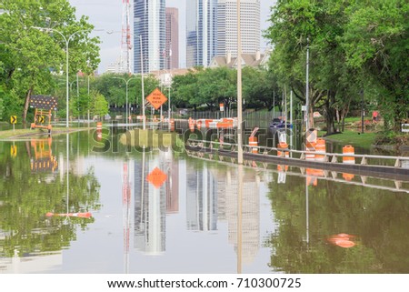 High water rising along Allen Parkway with road warning signs. Residential buildings and downtown Houston in background under cloudy sky. Heavy rains from tropical storm caused many floods. Swamp car