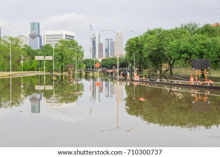 High water rising along Allen Parkway with road warning signs. Residential buildings and downtown Houston in background under cloudy sky. Heavy rains from tropical storm caused many floods. Swamp car