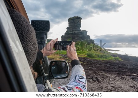 Man is taking picture of ancient rocks on mobile phone from the window of the car during safari off-road journey in Tundra of Kola Peninsula with Arctic Ocean on the background.