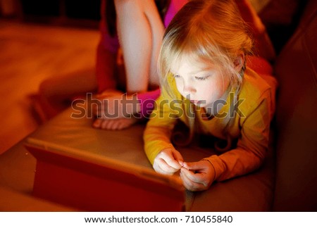 Adorable little girl playing with a digital tablet in a dark room. Children watching cartoons together at home. Education and learning for kids.
