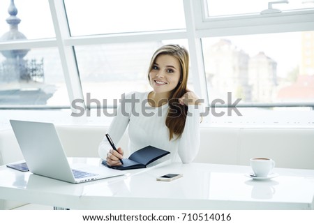 Portrait of cheerful smart student with notebook sitting at workplace with modern laptop computer in university.Positive female manager smiling at camera while writing down notes in notepad 