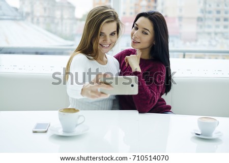 Two attractive females making selfie photo on modern smartphone for publication in social networks.Positive students taking pictures on front camera of digital cellular sitting in coffee shop