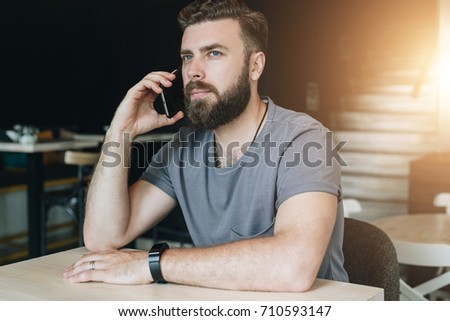 Portrait of young handsome bearded hipster man sitting in cafe at table and talking on his cell phone. Telephone conversations. Guy is calling his friends, girlfriend. Bokeh effect, film effect.