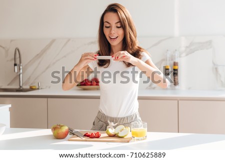 Happy smiling woman taking a picture of fruit slices on a wooden board with mobile phone while standing at the kitchen
