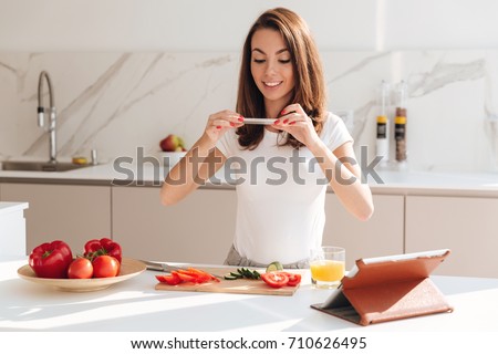 Smiling casual woman taking a picture of sliced vegetables on a wooden board with mobile phone while standing at the kitchen
