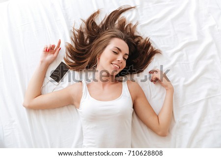 Top view of a smiling woman enjoys listening to music with earphones while laying on bed at home