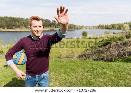 handsome smiling man holding ball and waving hand while looking away outdoors