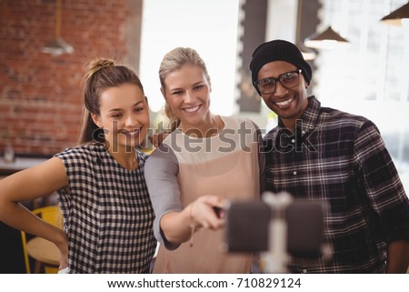 Smiling young woman taking selfie with friends from mobile phone on monopod at coffee shop