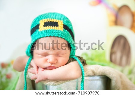 Newborn baby boy wearing a St. Patrick's Day hat sleeping in a silver metal bucket. Cartoon fairy house on background