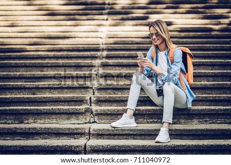 Attractive young female tourist is exploring new city. Woman with retro camera and mobile phone in hand is sitting on concrete stair. In search of new adventures.