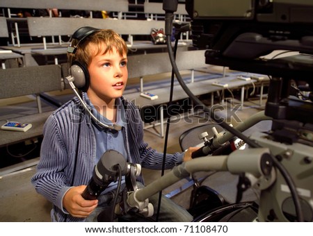 Little boy with headphones and microphone sits on the professional video camera in auditorium on television broadcast